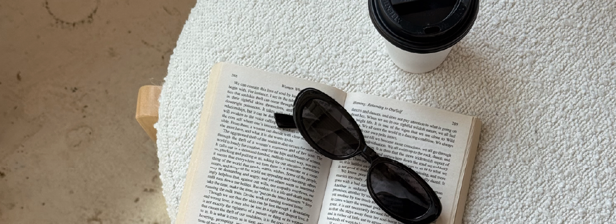 Open book with sunglasses on a cozy table, next to a takeaway coffee cup, set against a textured neutral background.
