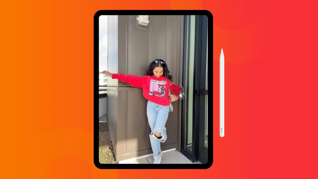 A girl in a red sweatshirt and ripped jeans poses playfully by a building, holding a bouquet of red roses. An iPad and pencil are adjacent.