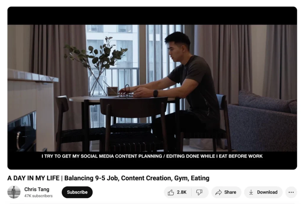 A person sits at a table, focused on a laptop, with a plant in a vase nearby and large windows providing natural light.