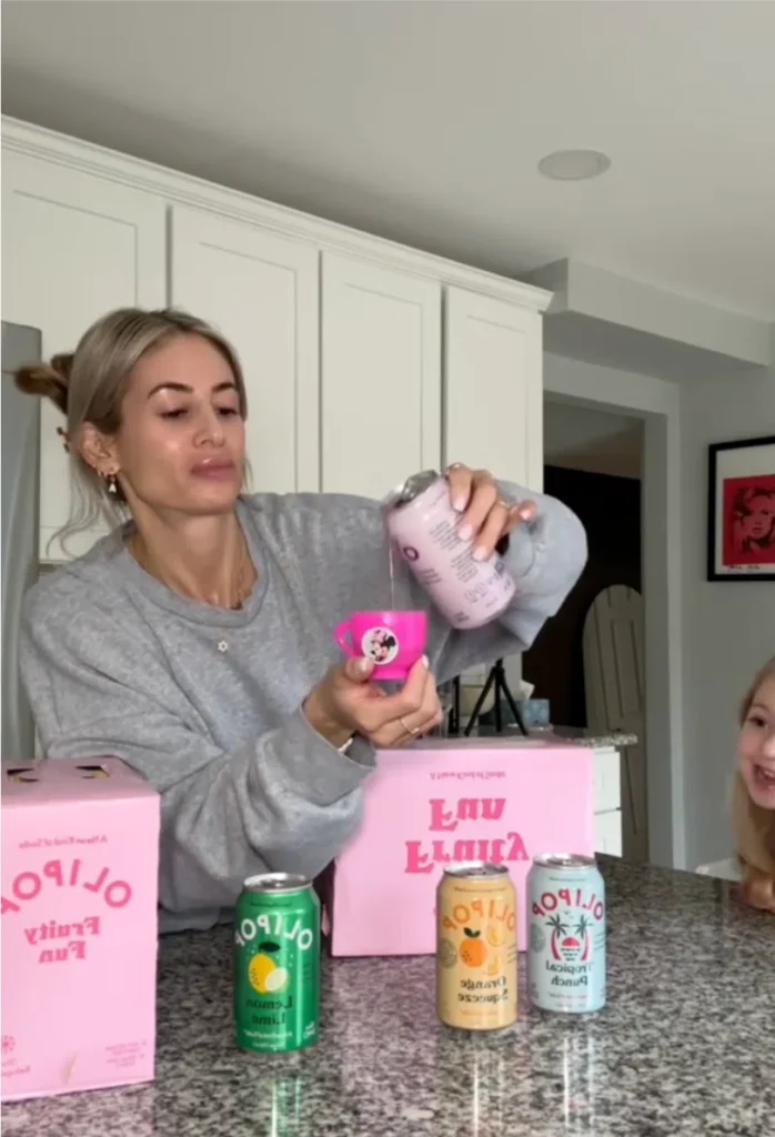 A person in a gray sweater pours a drink from a can into a pink cup while a child smiles nearby, with colorful cans on the counter.