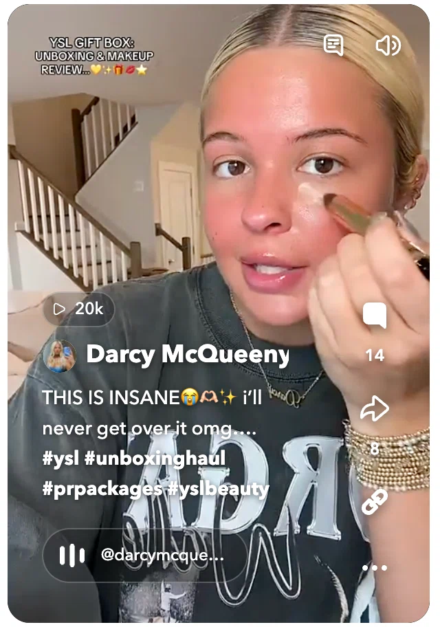 A woman wearing a graphic t-shirt gestures excitedly while unboxing a YSL gift box, showcasing her enthusiasm for makeup.