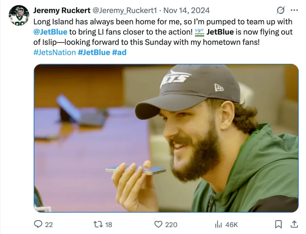A man in a green hoodie and Jets cap is focused on speaking into a phone, with a desk and blurred background visible.