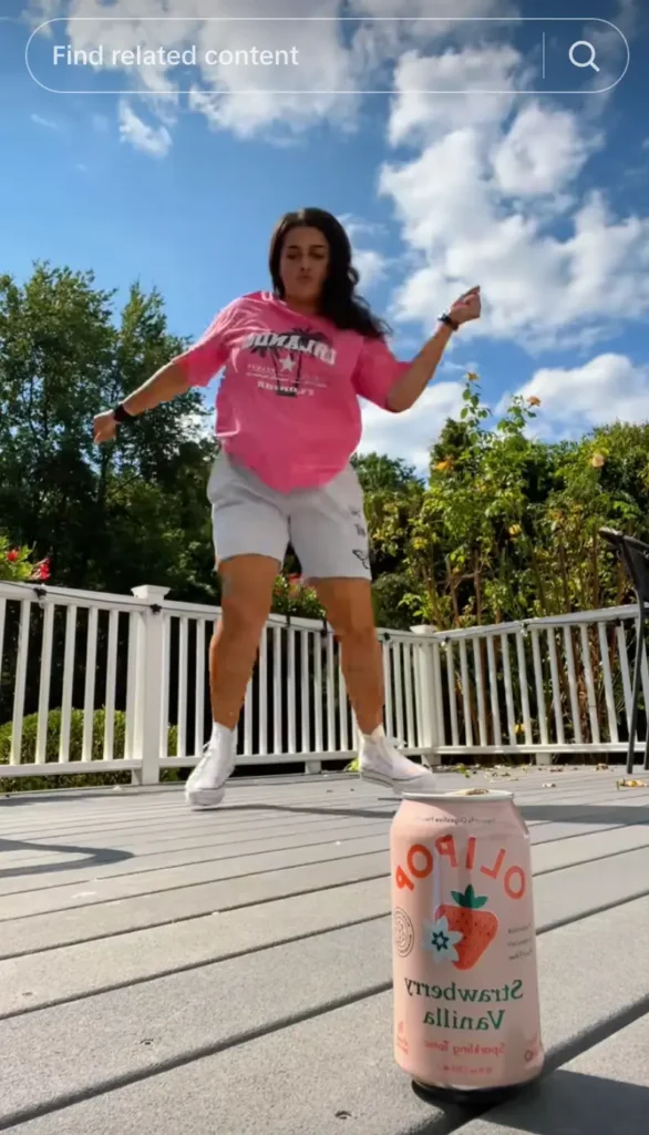 A person dances energetically on a deck, wearing a pink shirt and shorts, with a can of strawberry vanilla soda in the foreground.