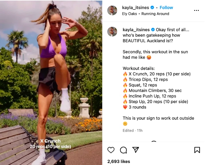 A woman exercises outdoors, performing an X Crunch on a bench, surrounded by lush greenery and colorful flowers. 