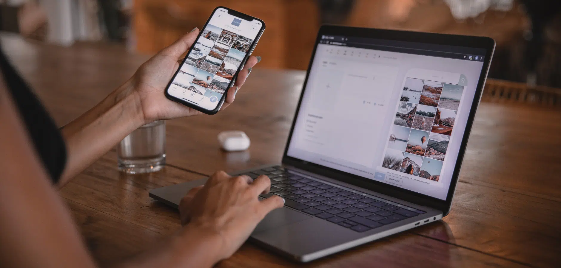 A person using a smartphone to view images while typing on a laptop, with a glass of water and earbuds on a wooden table.