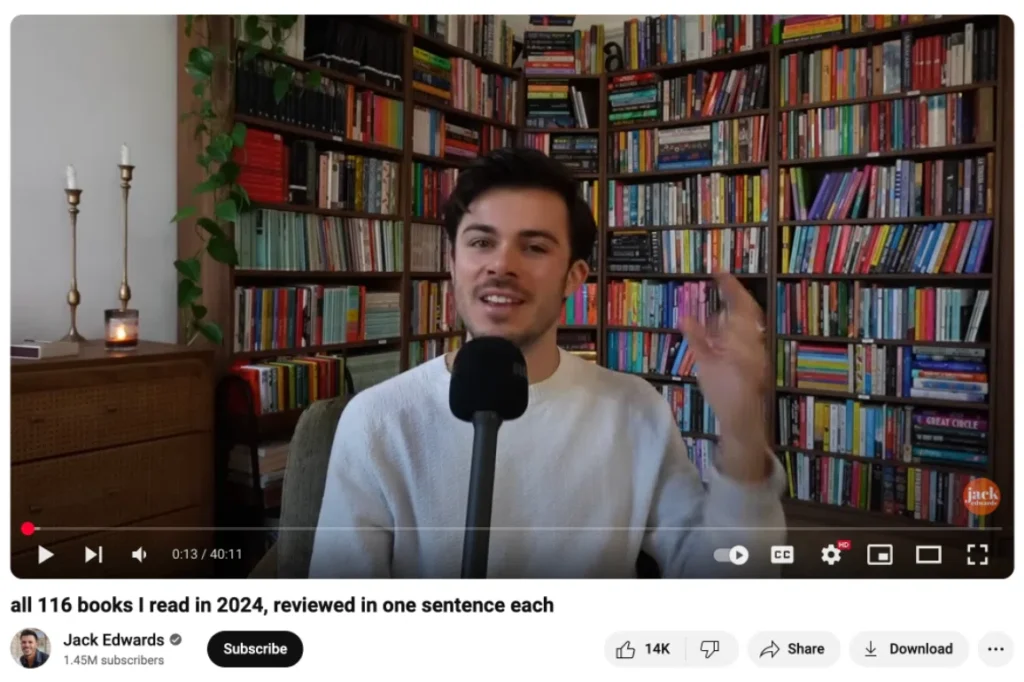 A person wearing a white sweater gestures while seated in front of a colorful bookshelf filled with various books.