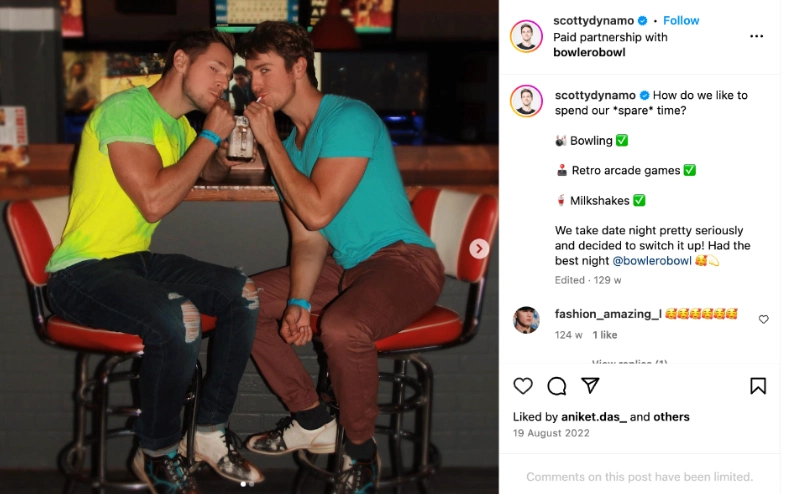 Two men sit close together at a brightly lit bar, sharing a milkshake with smiles, surrounded by retro arcade games and bowling decor.