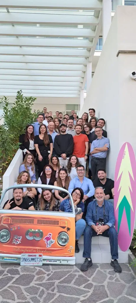 Group of people posing with a colorful van adorned with stickers and a surfboard on the side.