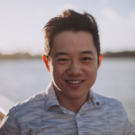 A person stands outdoors near a body of water, wearing a light gray shirt and smiling under a sunny sky with soft clouds.