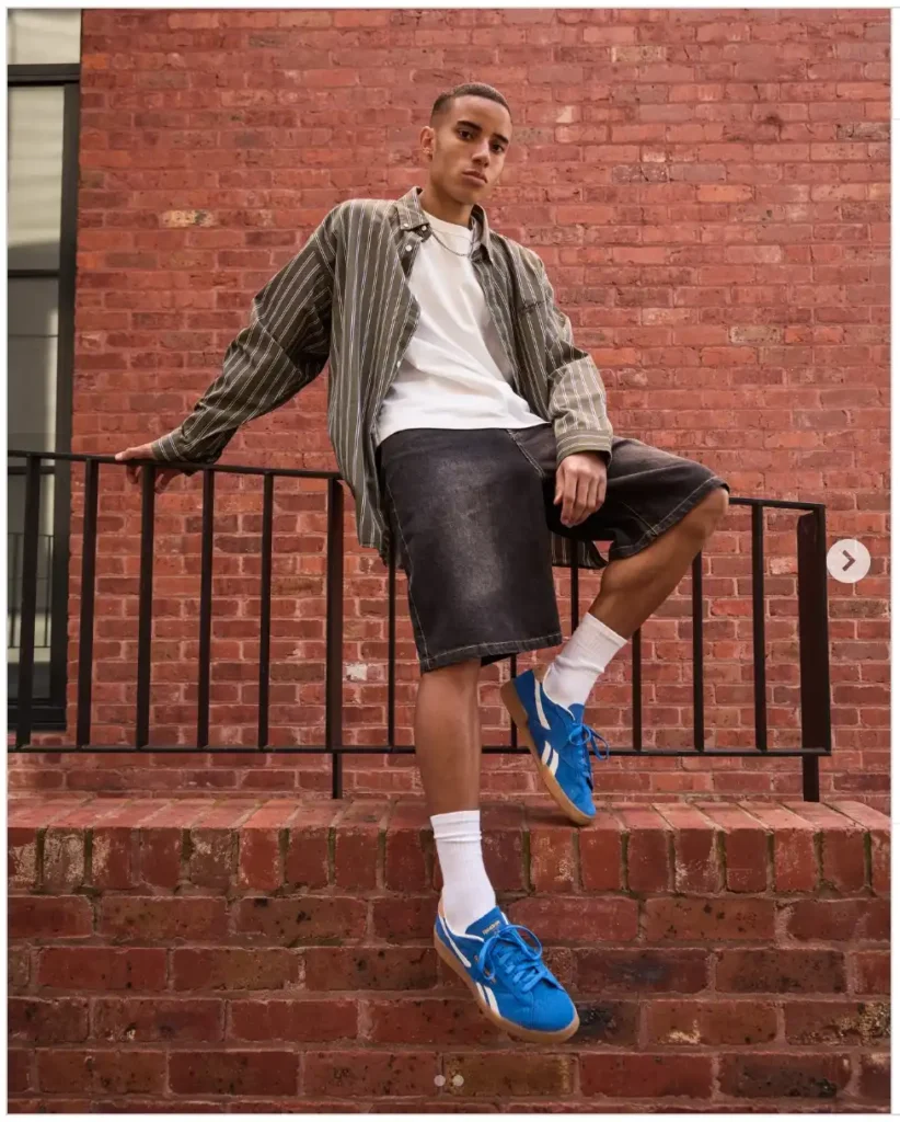 A person in a striped jacket and white tee leans against a railing, wearing blue sneakers and denim shorts, set against a brick wall.