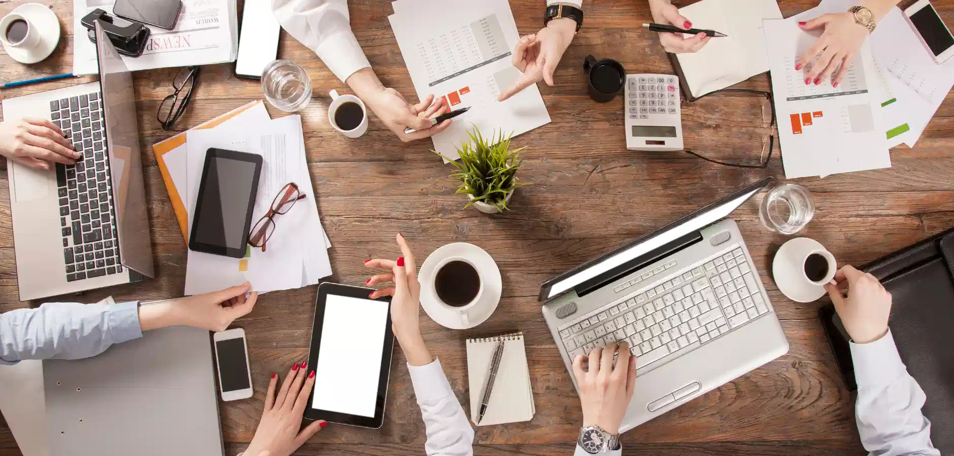 Overhead view of a busy workspace with hands holding devices, coffee, documents, and a laptop on a wooden table.