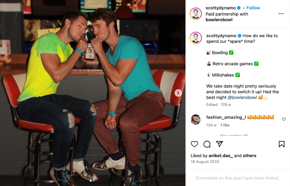 Two friends wearing brightly colored shirts toast with milkshakes at a bar, surrounded by retro arcade games and a lively atmosphere.