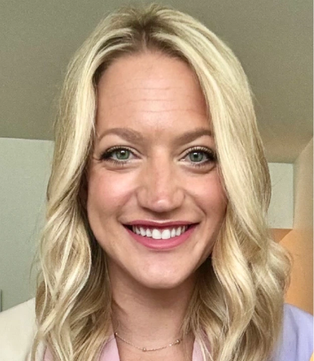 A person with wavy blonde hair wears a light-colored top and necklace, smiling against a softly blurred indoor background.