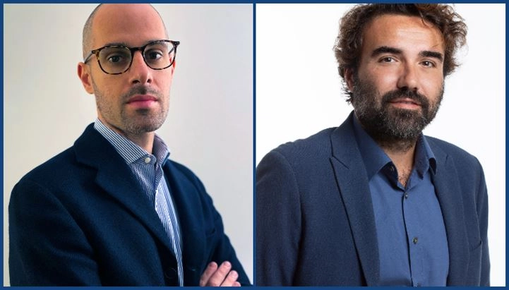 Two men in stylish jackets pose side by side. One has arms crossed; the other stands with hands at his sides, both in professional attire.