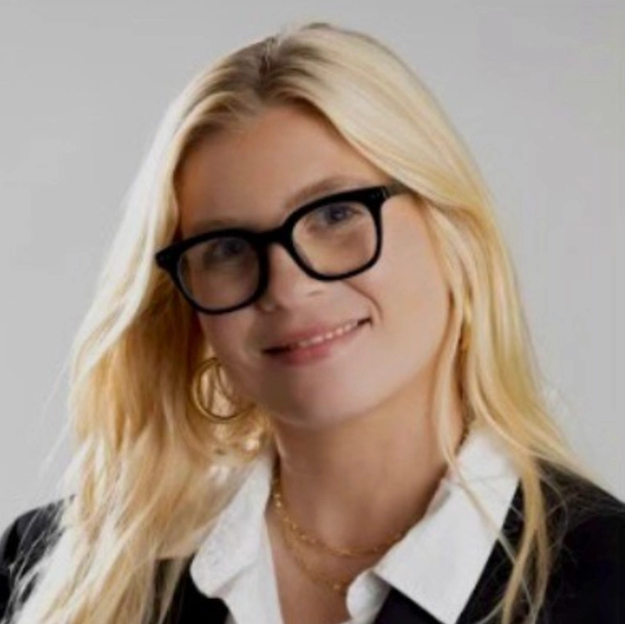 A person with long blonde hair wearing a black blazer over a white shirt, set against a light gray background.