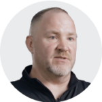 A man wearing a black zip-up shirt stands against a plain background, captured in a circular crop.