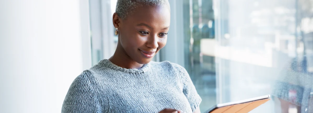 A person stands in front of a large window, intently using a tablet, wearing a cozy knit sweater and small earrings.