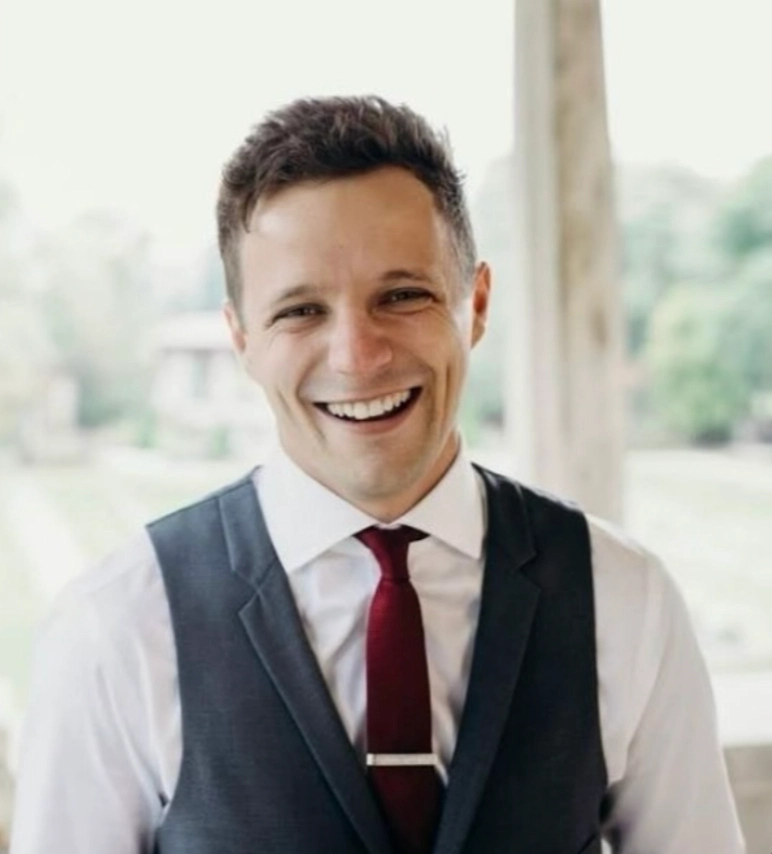 A man in a formal outfit with a dark vest and white shirt, wearing a red tie, stands in a blurred outdoor setting.