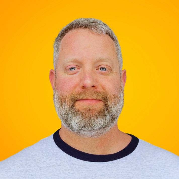 A person with short gray hair and a beard wearing a gray t-shirt with a dark collar against a bright yellow background.