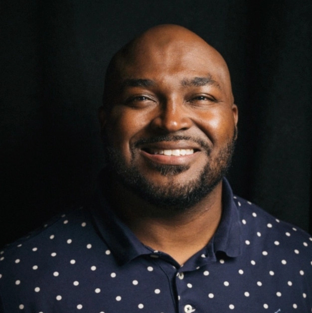 A person wearing a navy blue polka dot shirt stands against a dark backdrop, exuding a serious and calm demeanor.