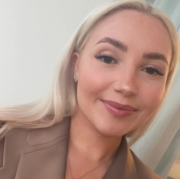 A person with long, sleek blonde hair wears a stylish brown blazer and elegant earrings, posing in a softly lit room.