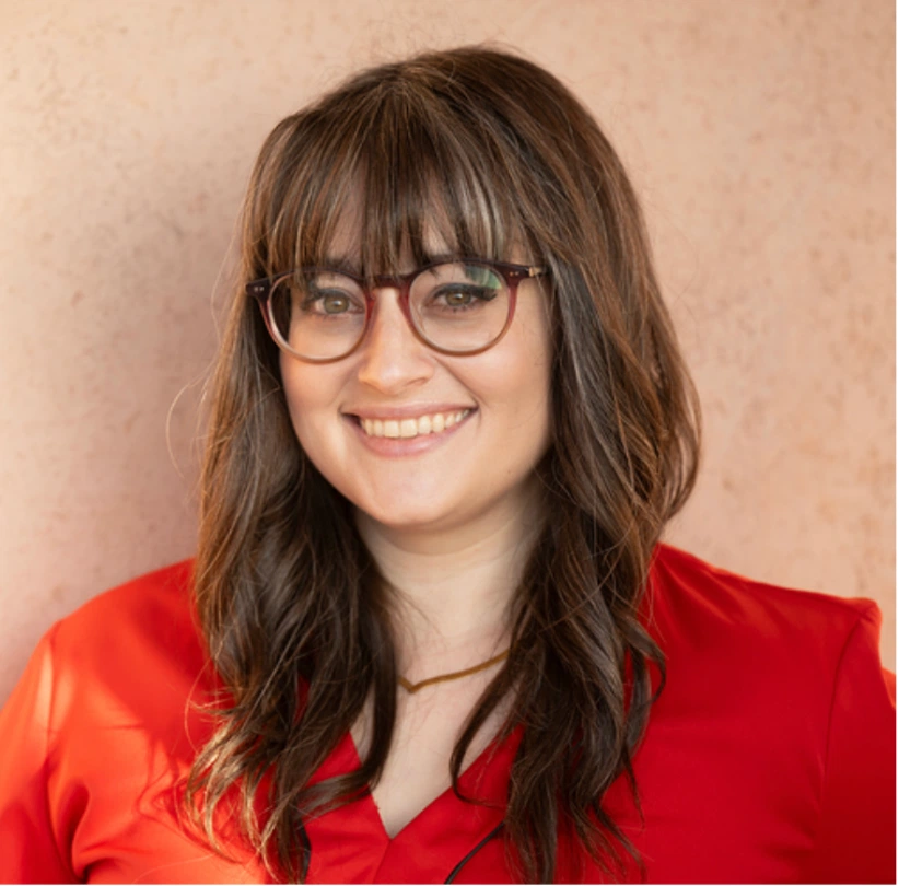 A woman in a vibrant red blouse stands against a soft, textured pink background, showcasing long, wavy brown hair.