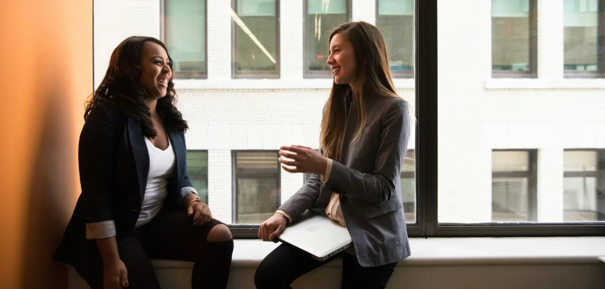 Two professionals engage in a discussion by a large window, overlooking a cityscape, with one holding a laptop.