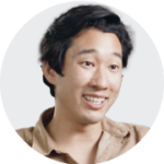 A close-up portrait of a person with dark, wavy hair, wearing a light brown shirt against a plain white background.