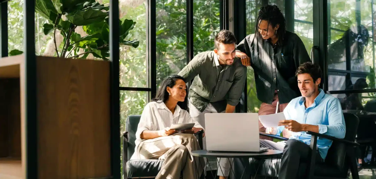 A diverse group of professionals collaborates in a modern workspace with plants and a laptop, discussing ideas and reviewing documents.