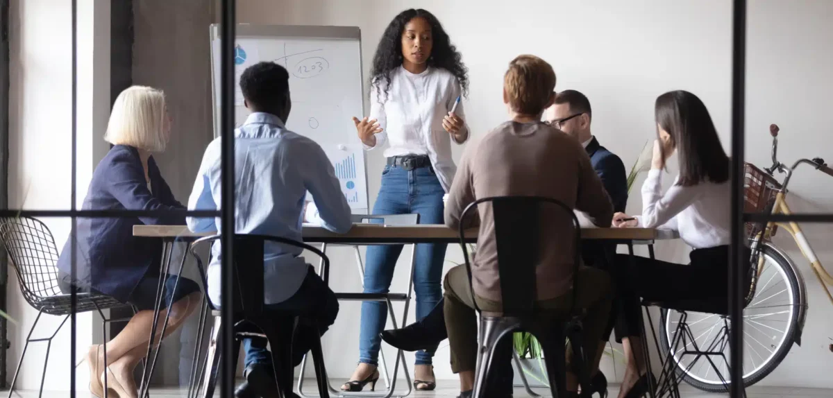 A diverse group of individuals in a meeting, attentively listening to a speaker at the front, with a bicycle visible in the background.