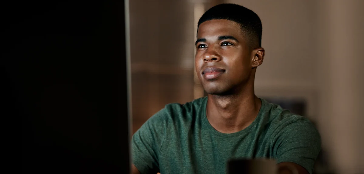 A person in a green shirt sits at a computer, focused on the screen in a softly lit room.