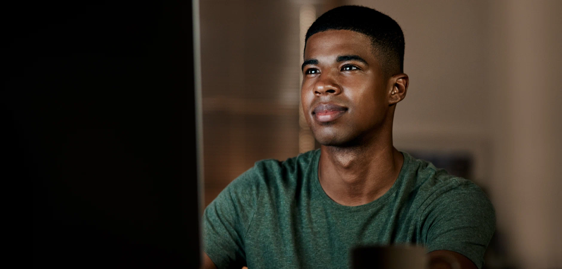 A person in a green shirt sits at a computer, focused on the screen in a softly lit room.