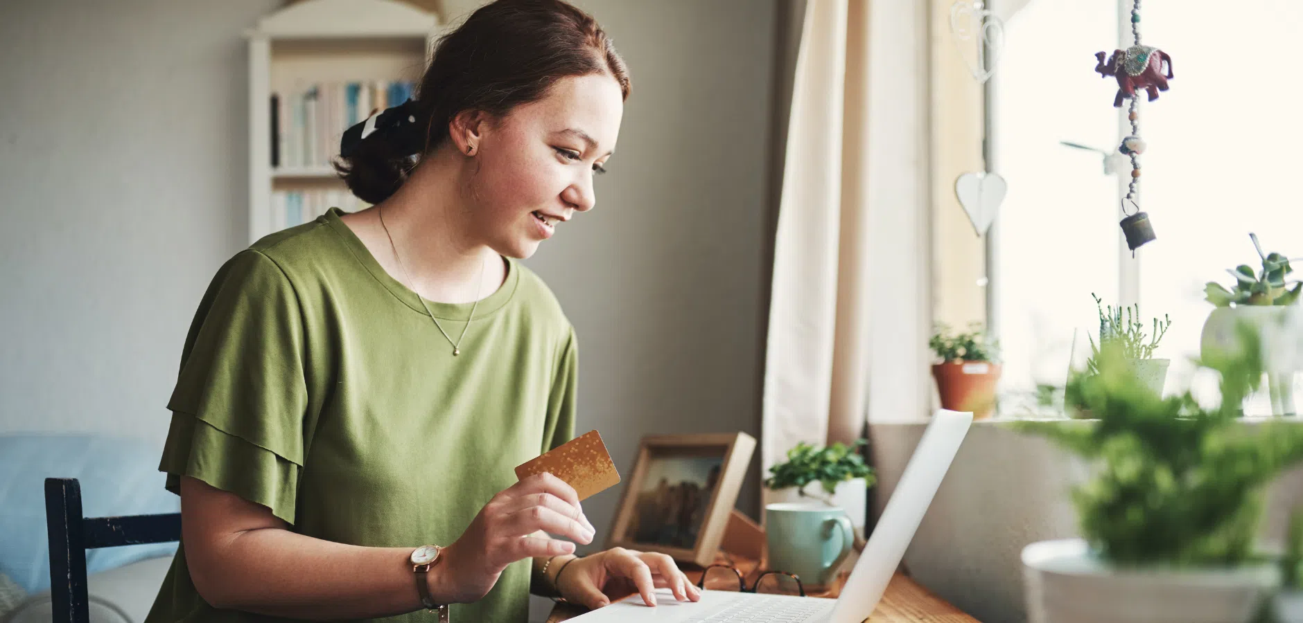 A woman in a green shirt holds a credit card, focused on her laptop in a cozy room with plants and natural light.