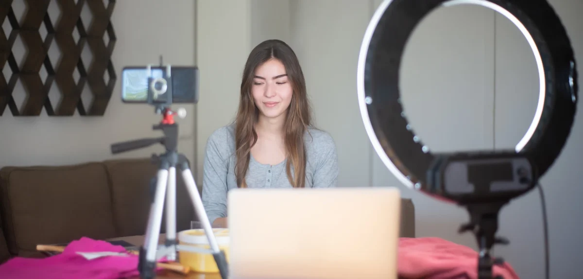 A person sits in front of a laptop and a ring light, filming content for social media in a cozy indoor setting with colorful props.