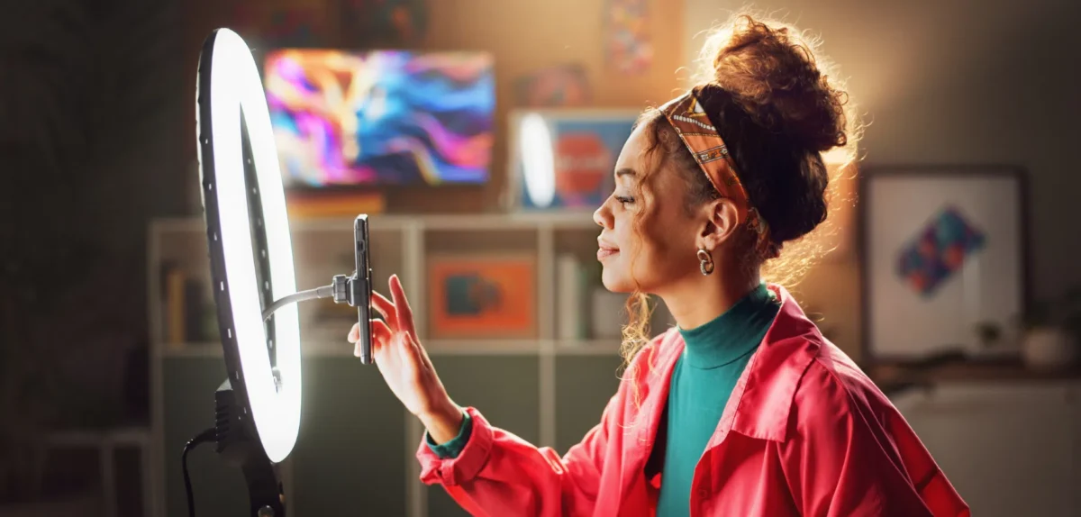 A woman in a pink jacket is posing in front of a ring light, holding a smartphone mounted on a stand in a well-lit room.