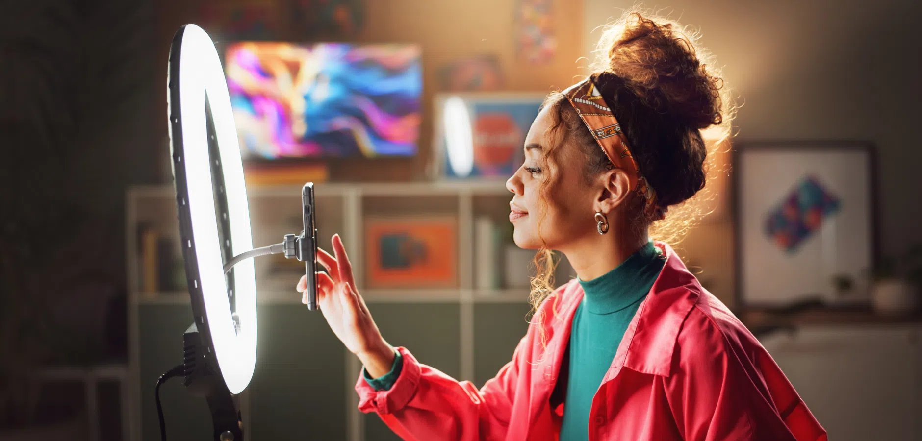 A woman in a pink jacket is posing in front of a ring light, holding a smartphone mounted on a stand in a well-lit room.