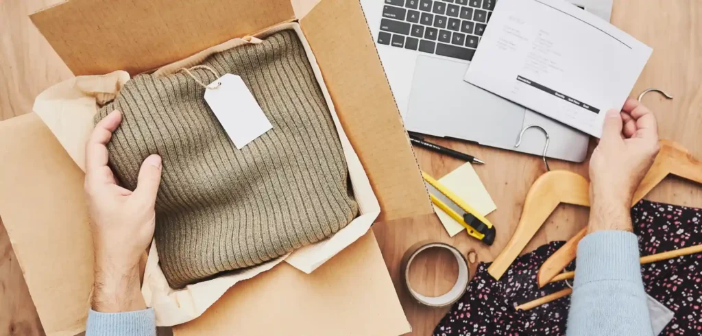 A person unboxes a ribbed olive sweater from a cardboard box, surrounded by a laptop, documents, and clothing hangers.