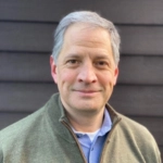 A person wearing a green sweater over a light blue shirt stands in front of a dark wooden background.