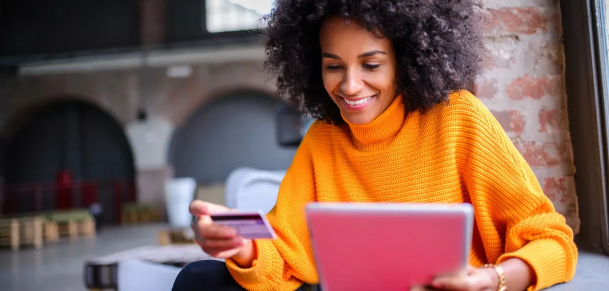 A person in an orange sweater holds a credit card in one hand and a pink tablet in the other, seated in a modern, industrial space.