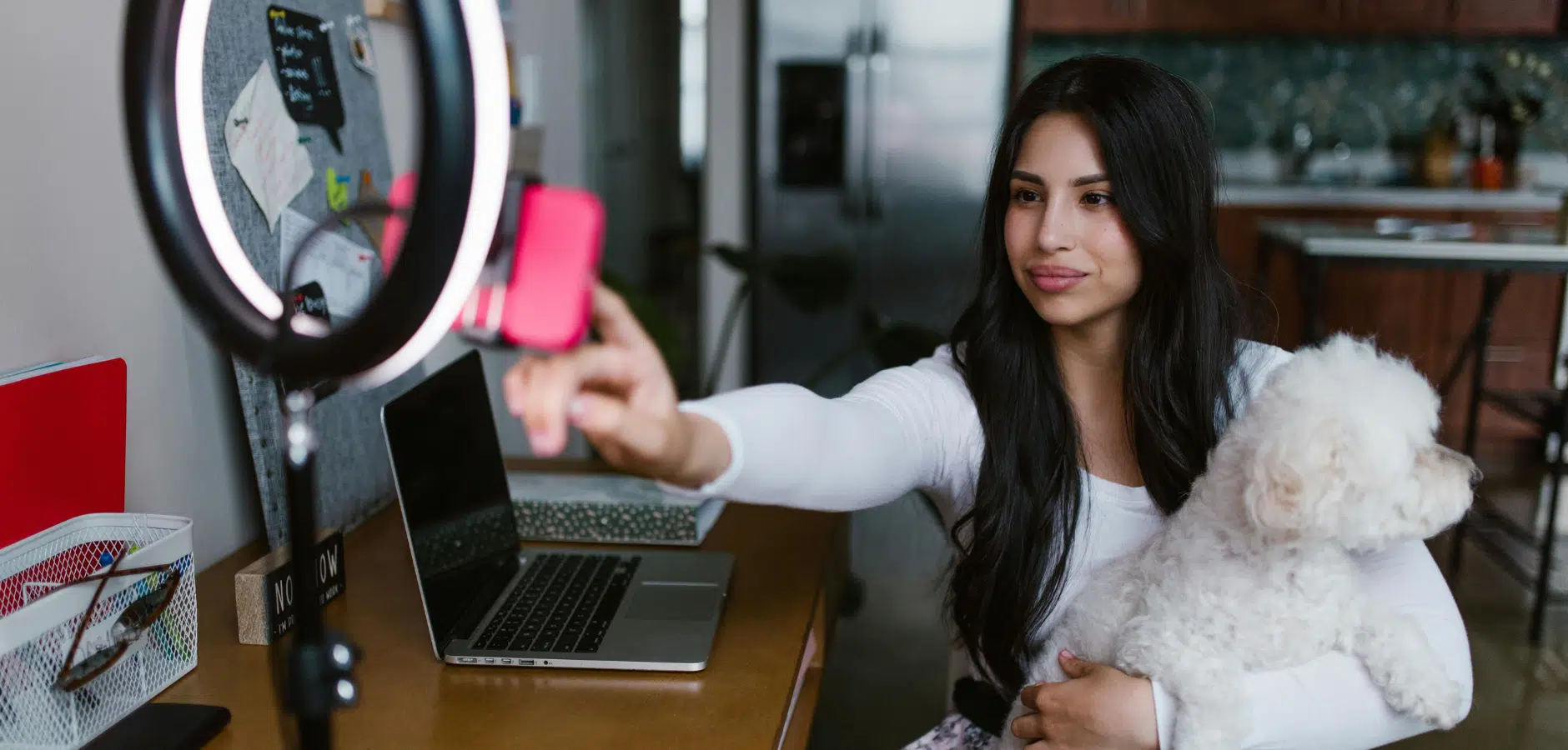 A woman sits at a desk with a laptop and a ring light, holding a small white dog in her arms, pointing towards the camera.