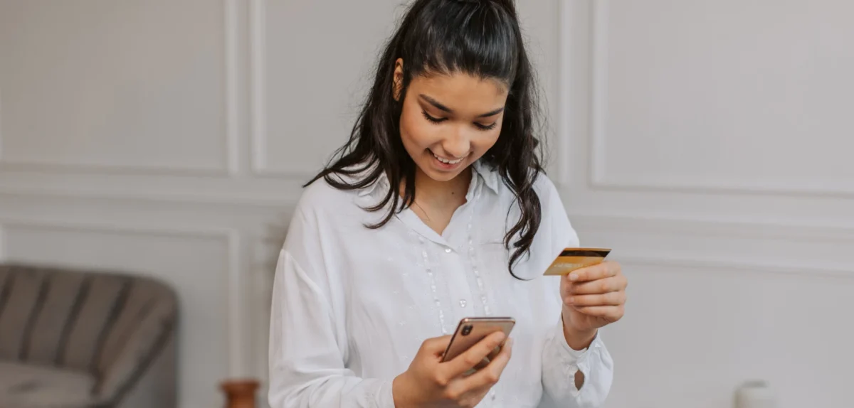 A person in a white shirt holds a phone in one hand and a credit card in the other, preparing for an online transaction.