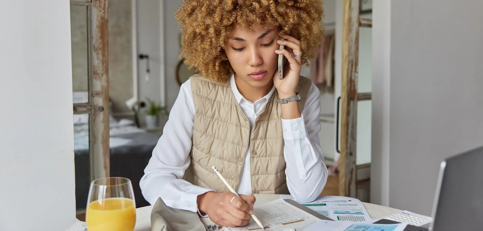A person in a beige vest and white shirt speaks on the phone, surrounded by documents and a glass of orange juice on a table.