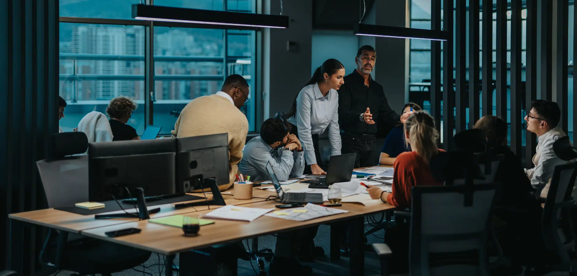 A diverse group of professionals engaged in a meeting surrounded by laptops and documents in a modern office setting with city views.