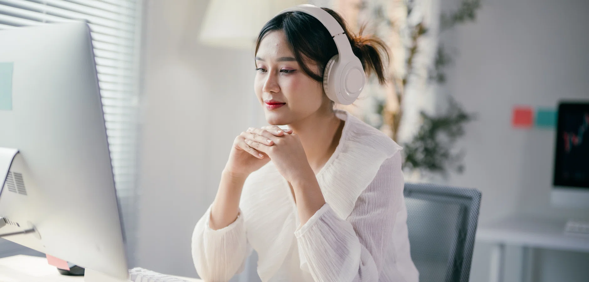 A person wearing headphones sits in front of a computer, hands clasped thoughtfully, in a bright, modern workspace.