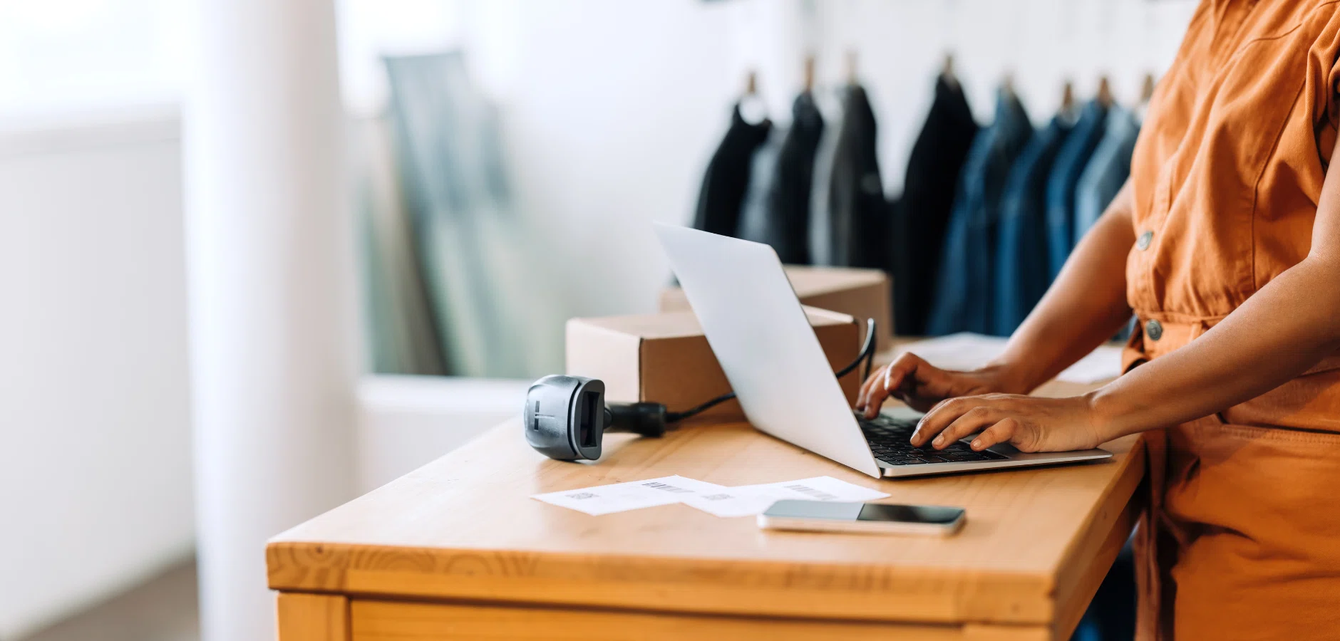 A person in a brown jumpsuit types on a laptop at a wooden desk, surrounded by shipping boxes and a barcode scanner.
