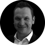 A professional individual wearing a black suit with a white shirt, set against a solid black background.