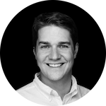 A man with short, light brown hair wears a white collared shirt against a solid black background.
