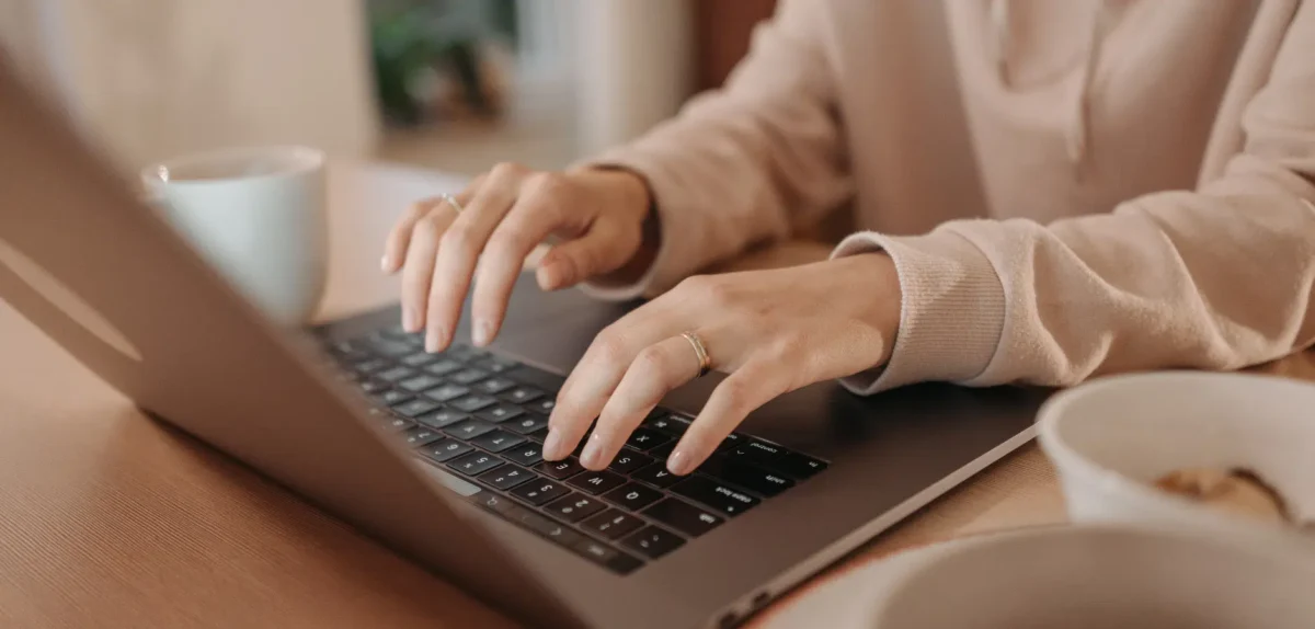 A person in a cozy sweatshirt types on a laptop, with a mug and bowl of food on a wooden table nearby.
