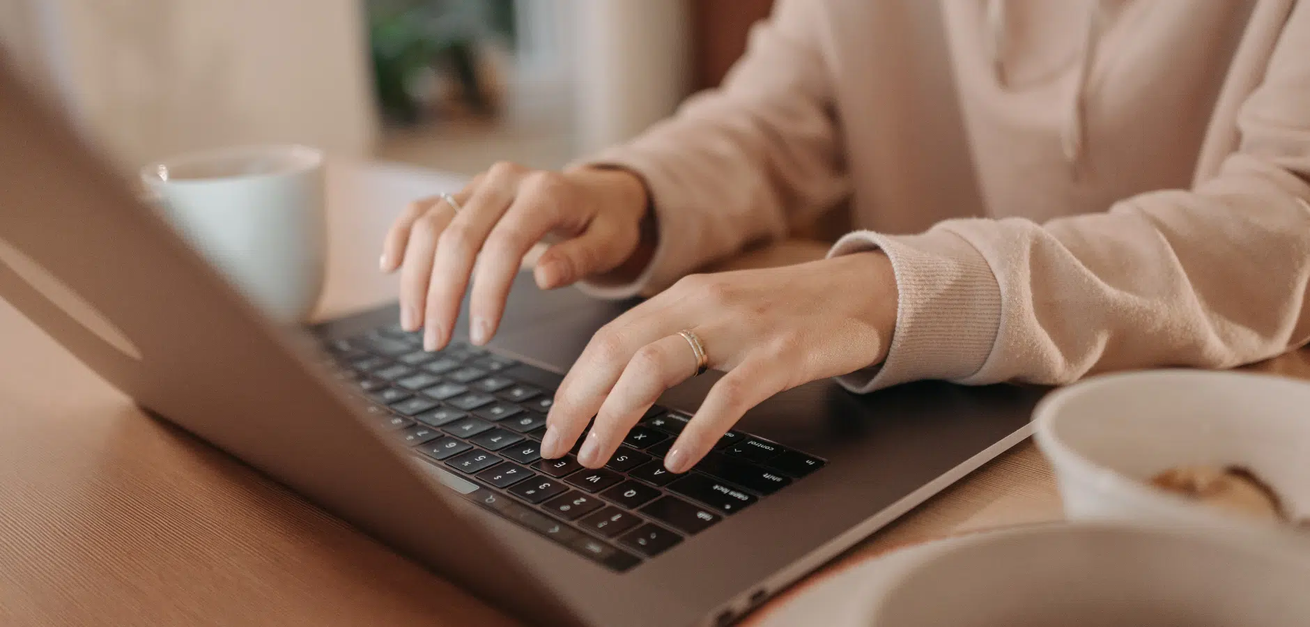 A person in a cozy sweatshirt types on a laptop, with a mug and bowl of food on a wooden table nearby.