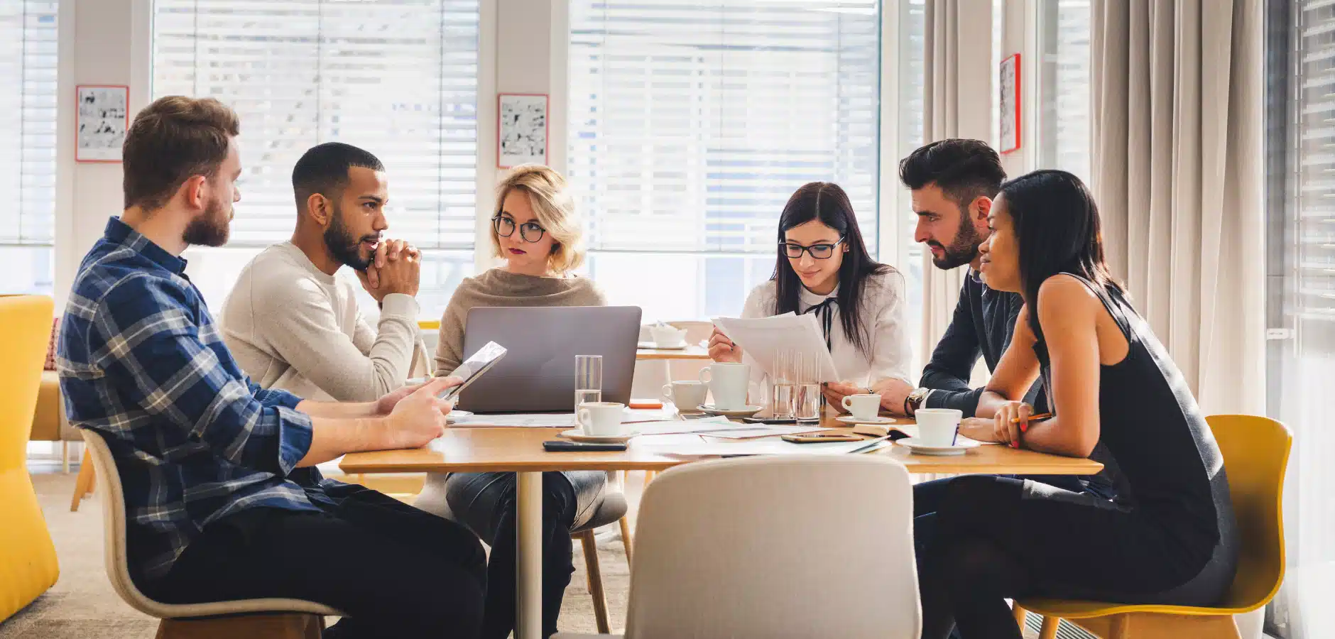 A diverse group of six people sits around a table, discussing documents and using laptops, with coffee cups scattered around.